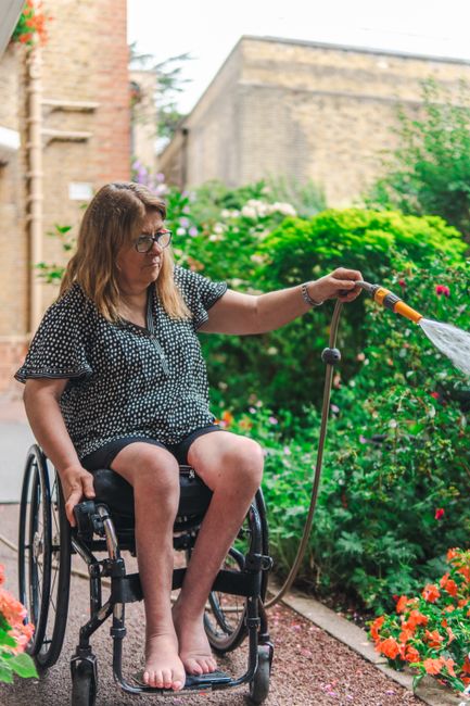 A woman in a wheelchair, wearing glasses and a black and white polka-dotted dress, sits in a garden with vibrant flower beds and lush green shrubs. She is holding a hose and watering the plants, with 